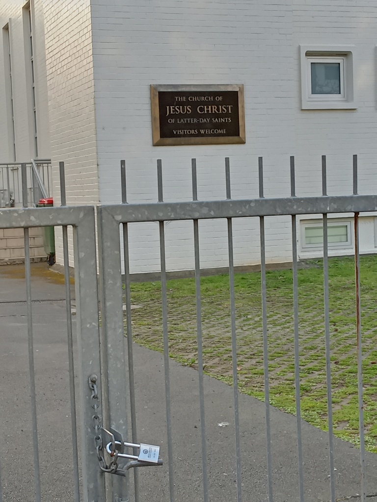 A photo. Behind a locked and chained gate the sign says "The Church of Jesus Christ of Latter-Day Saints. Visitors Welcome"